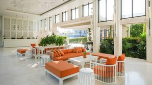 Un vestíbulo espacioso con diseño moderno, que cuenta con un amplio espacio de descanso con mobiliario naranja y blanco, una recepción y grandes ventanales con vistas al exterior.