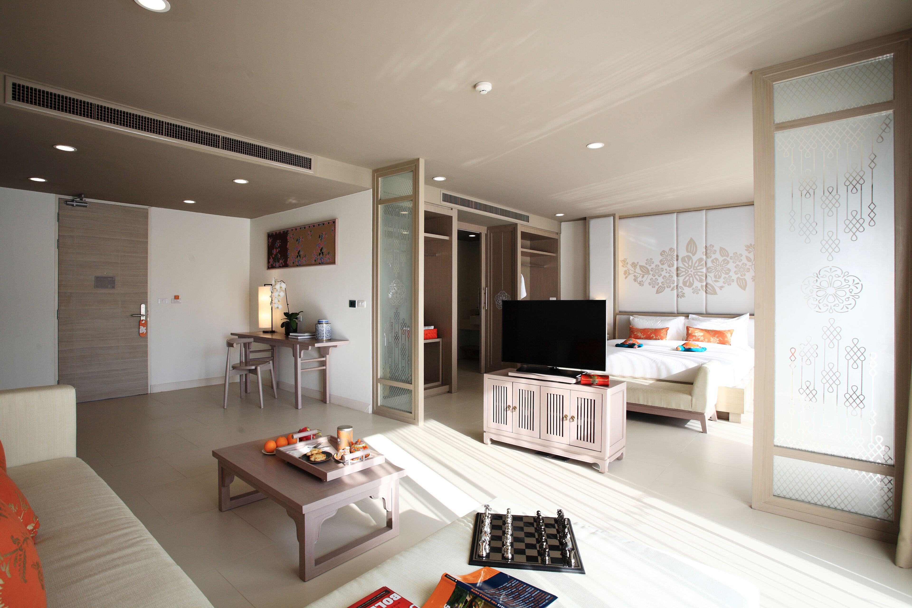 A modern hotel room with a flat-screen TV, a bed with decorative pillows, a dining area with a table and chairs, and a closet with sliding doors.