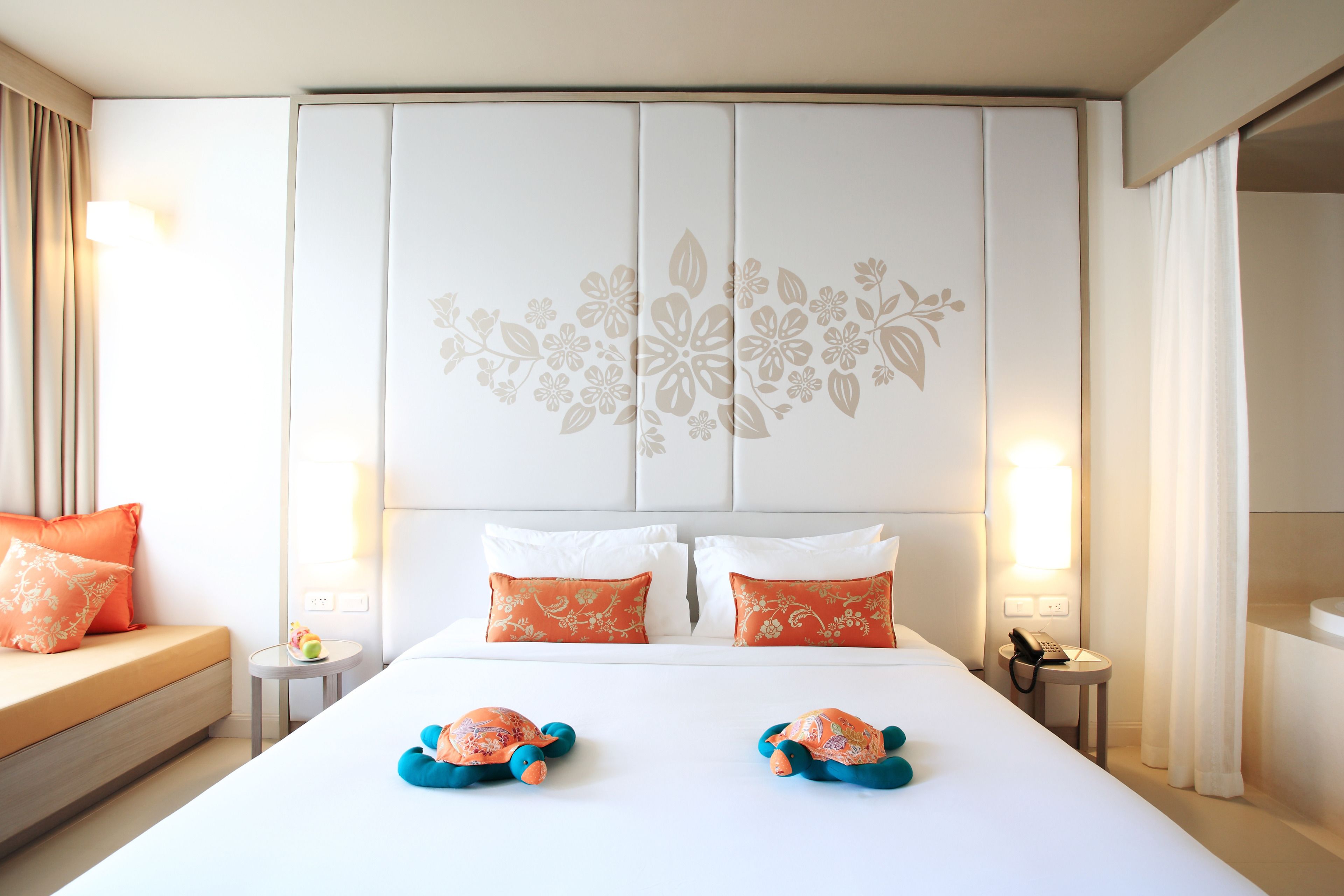 Une chambre d’hôtel avec un grand lit, deux coussins décoratifs, une table de chevet avec une lampe et une petite table avec un jouet.