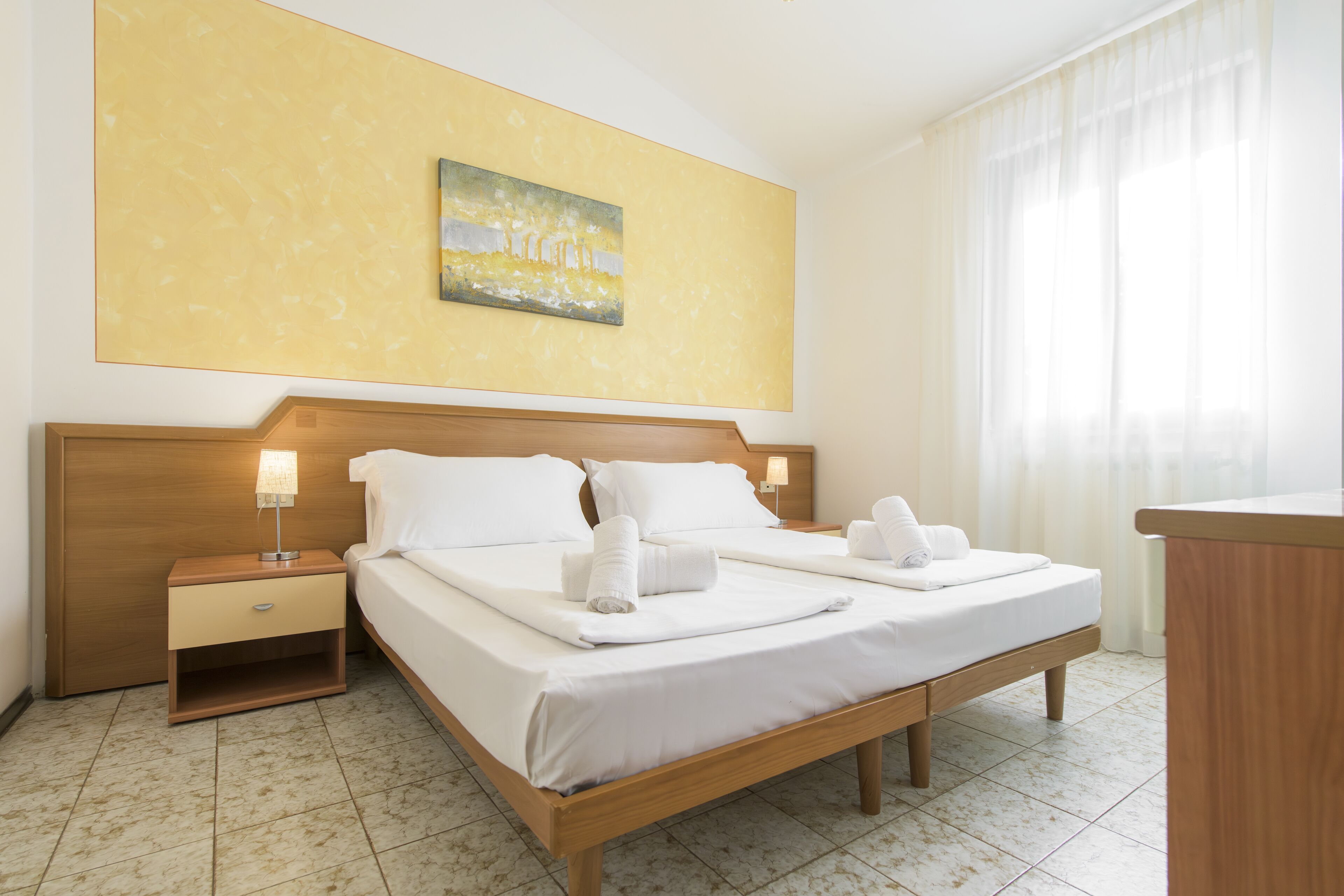 Habitación de hotel con cama doble, mesita de noche con lámpara, un cuadro en la pared y ventana con cortinas.