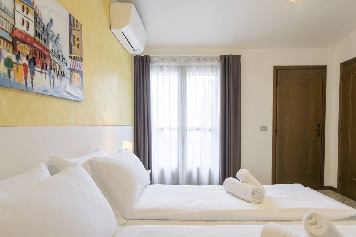 A hotel room with a large bed, a painting on the wall, a window with curtains, and a door.