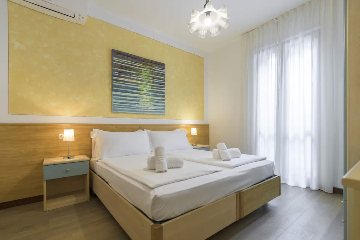 A double bed with white linens, two bedside tables with lamps, a large abstract painting on the wall, and a large window with sheer curtains.