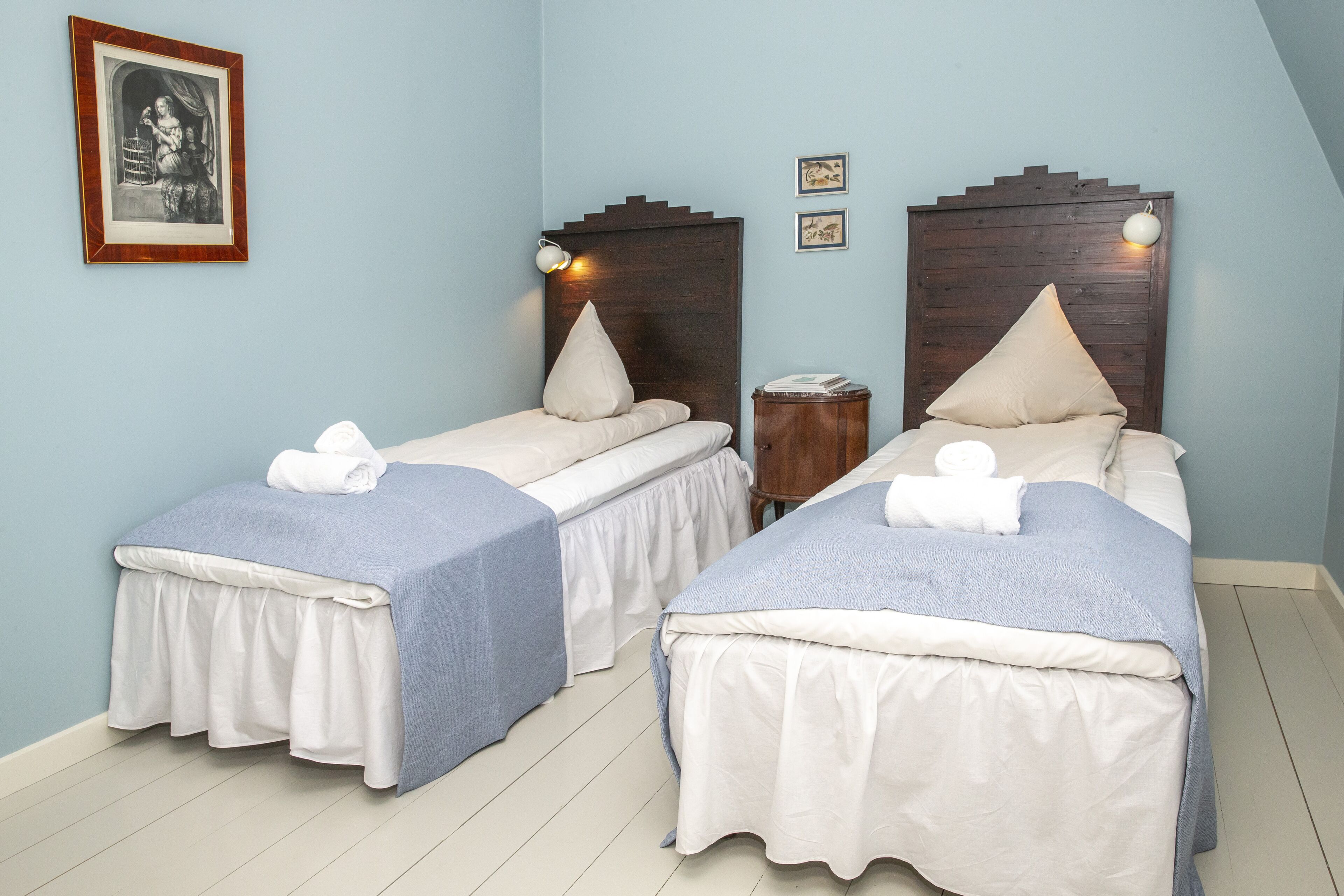 A room with two single beds, each with a wooden headboard, white bedding, and a blue skirt. There is a small wooden nightstand with a lamp between the beds.