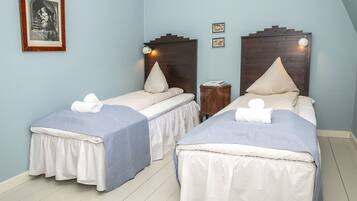 A room with two single beds, each with a wooden headboard, white bedding, and a blue skirt. There is a small wooden nightstand with a lamp between the beds.