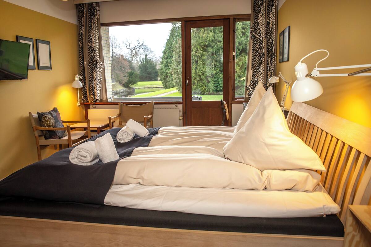 A neatly made bed with pillows and a rolled towel, a wooden headboard, a bedside table with a lamp, and a window with a view of a garden.