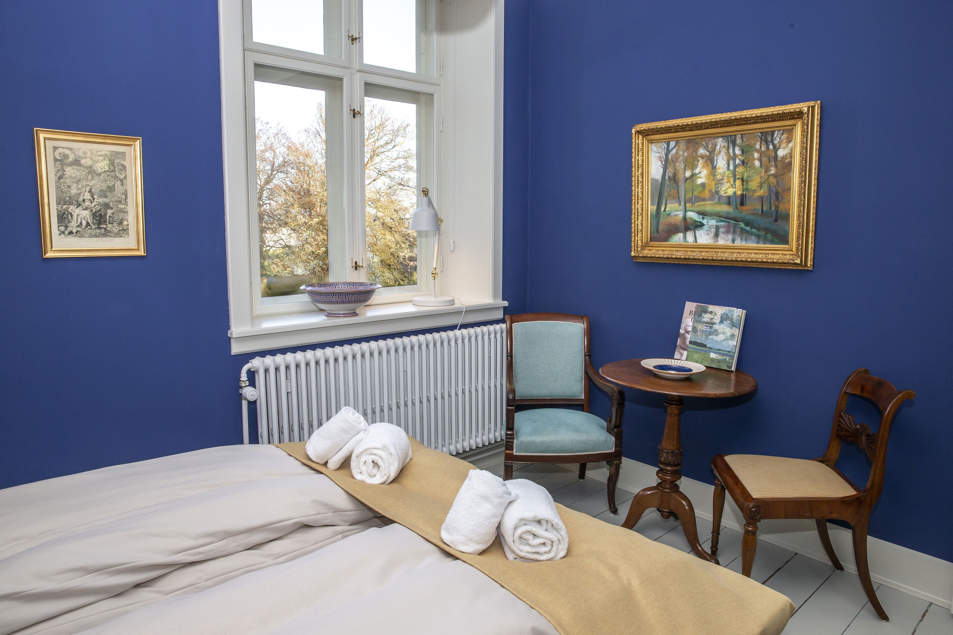 A hotel room with a bed, a table with a bowl, two chairs, and a window with a view of trees.