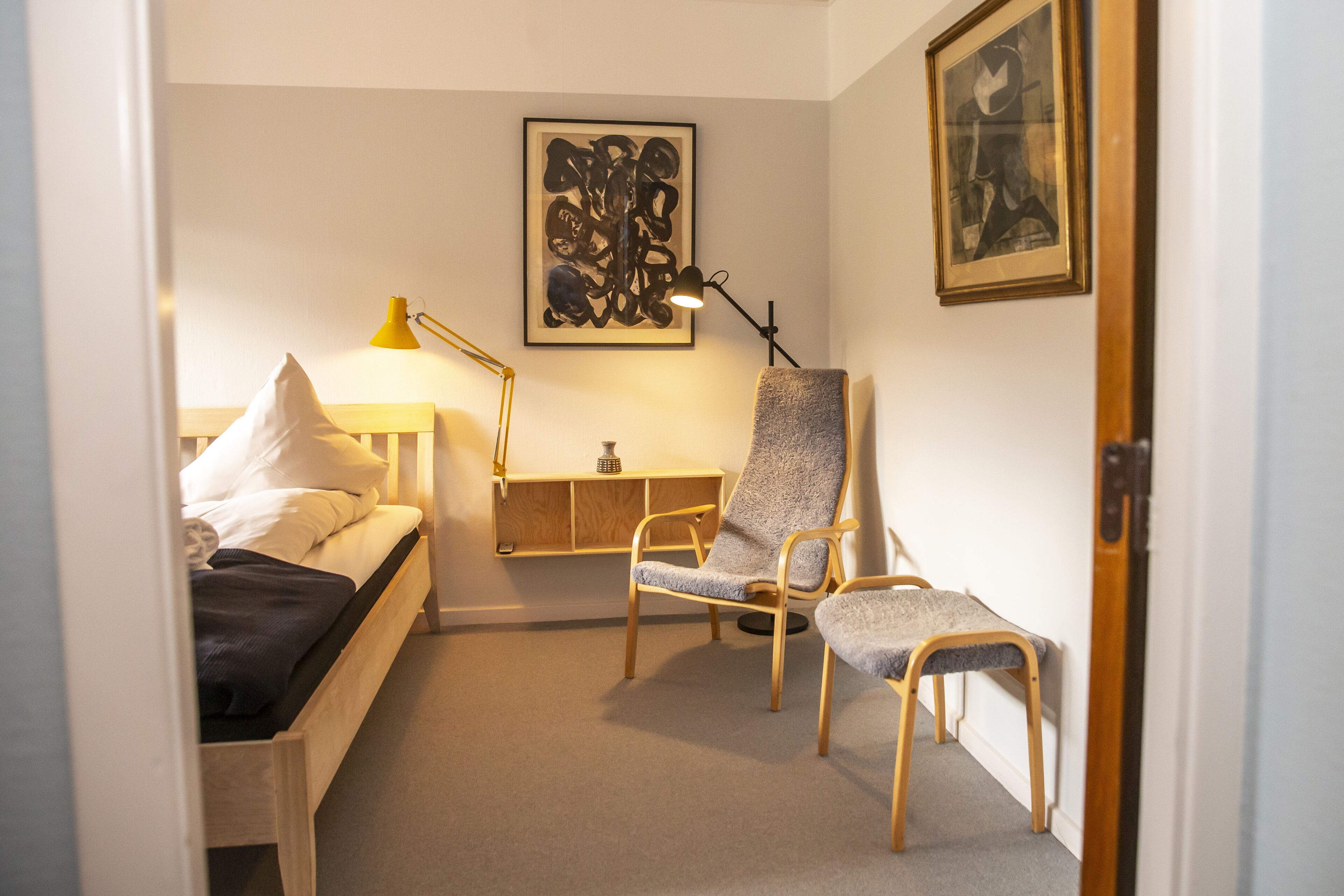 A bedroom with a bed, two chairs, a desk, a lamp, and artwork on the walls.