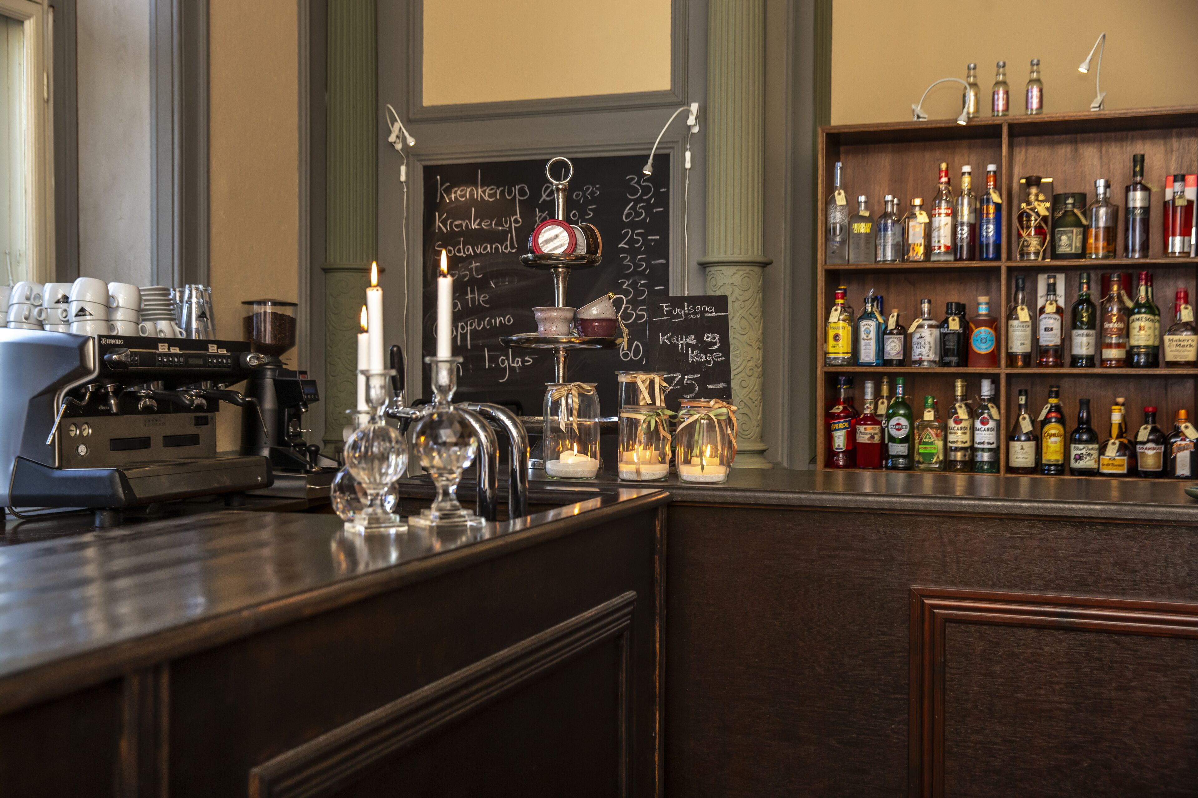 Uma área de bar com uma máquina de café, um quadro negro com o cardápio e uma estante com garrafas de bebidas.