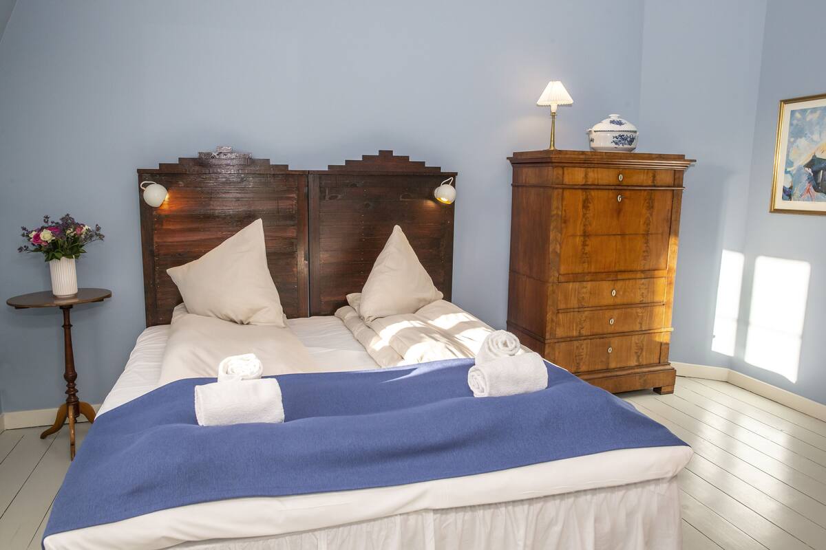 A neatly made bed with a blue blanket, white pillows, and two towels. A wooden dresser with a lamp and a vase of flowers is beside the bed.
