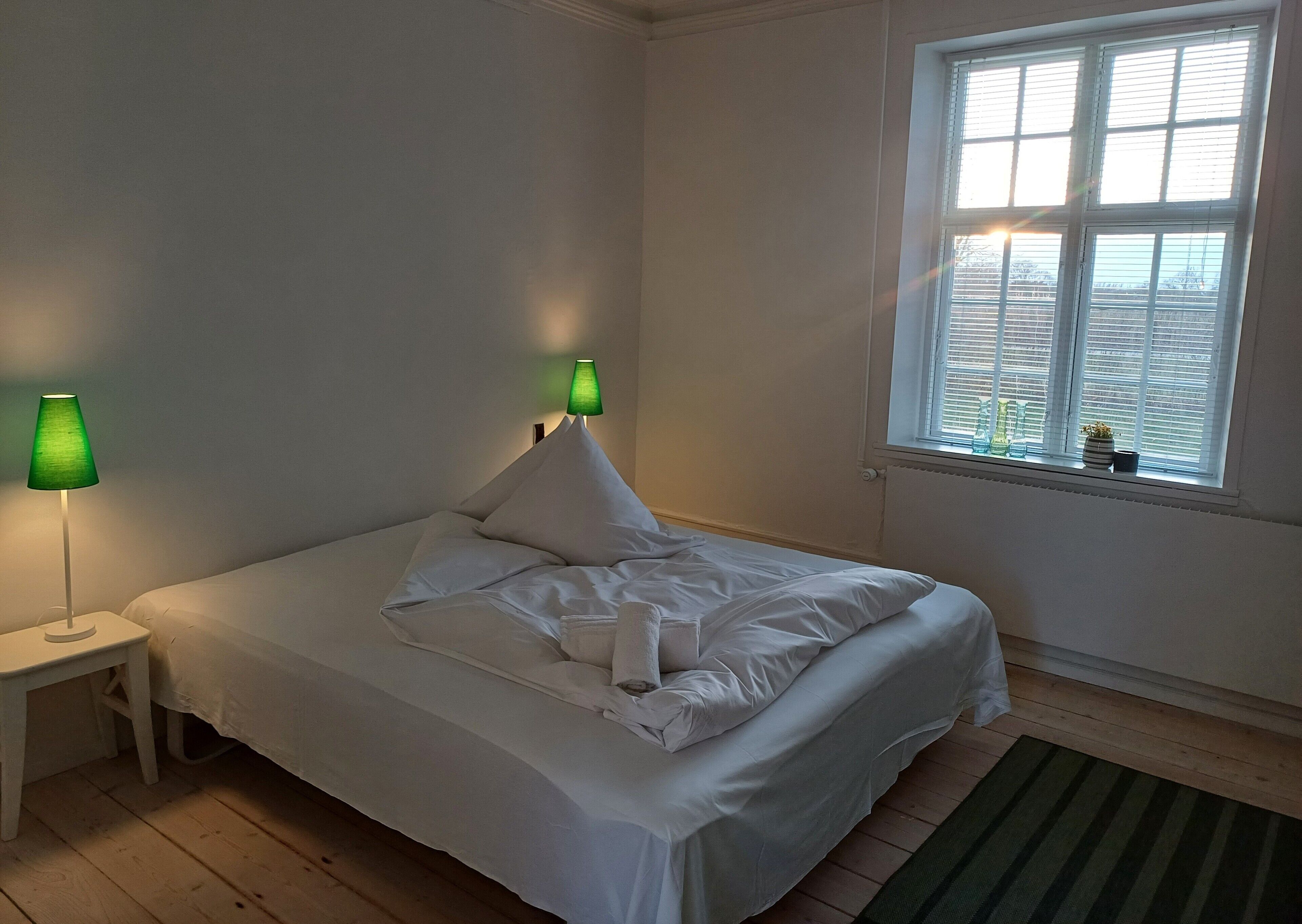 A neatly made bed with white linens, two bedside lamps, a window with blinds, and a small plant on the windowsill.