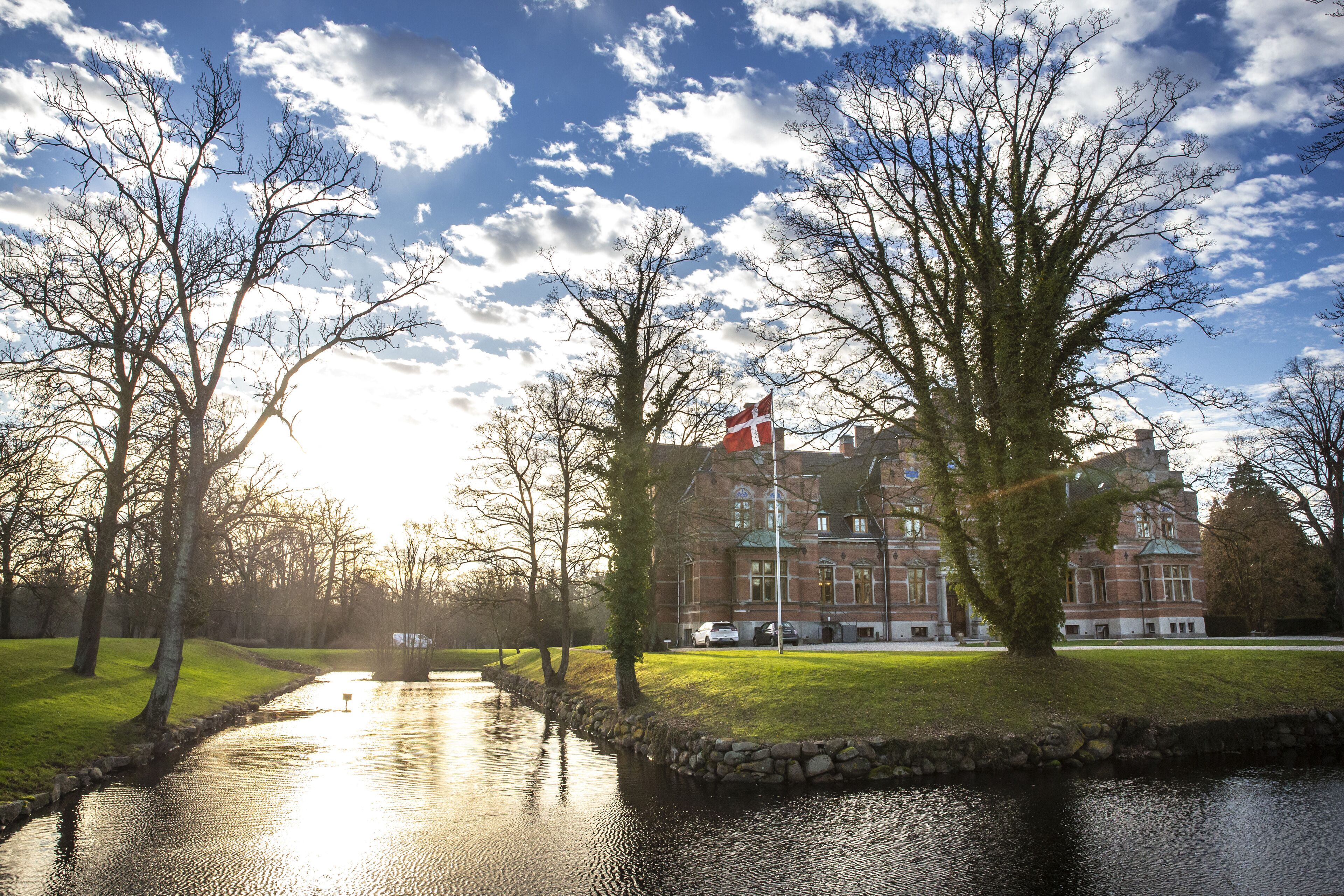 A riverside view with a building featuring a flag, stone-lined banks, and trees.