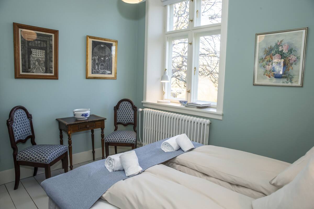 A room with a bed, two chairs, a table with a bowl, and framed pictures on the wall.