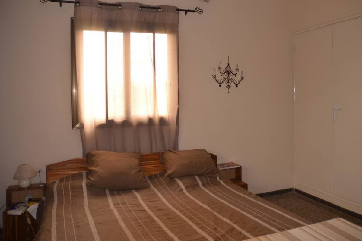 A bedroom with a bed, curtains, a nightstand, a lamp, a wall-mounted light fixture, and a wardrobe.