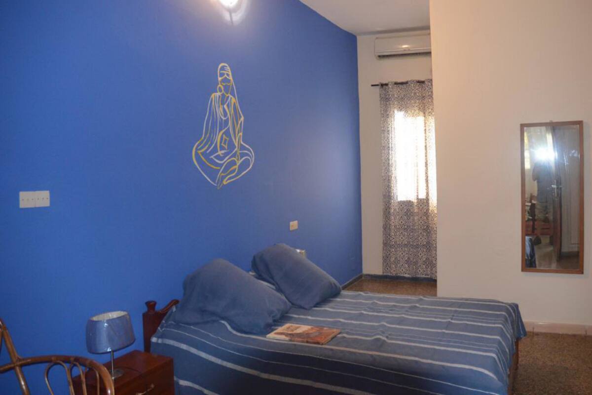 A bedroom with a bed, a chair, a lamp, and a wall with a decorative line drawing.