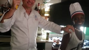 A chef holding a spatula and a pot, standing in a kitchen with various cooking utensils and bottles in the background. - Hotel Restaurant Coté Sud (Lomé)