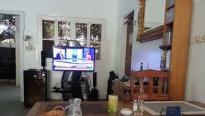 A dining area with a television displaying a news program, a coffee table with a basket, pitcher, and plates, and a bookshelf with books and a mirror. - Hotel Restaurant Coté Sud (Lomé)
