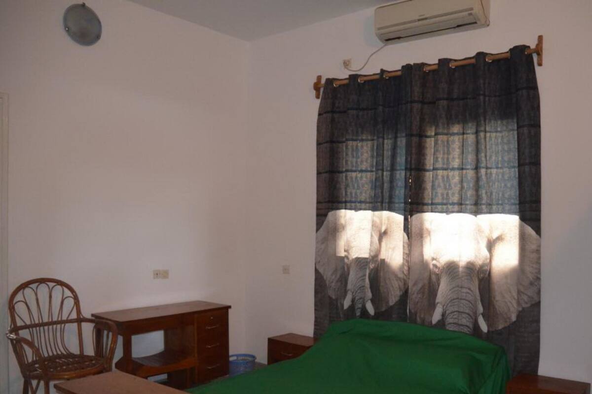 A small, simple room with a bed, a desk, a chair, and an air conditioning unit.