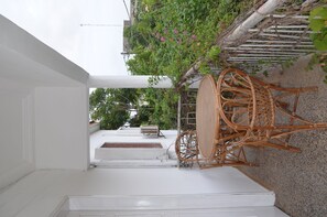A white outdoor area with a wicker table and chairs, a white wall, and a small garden with green foliage and pink flowers.