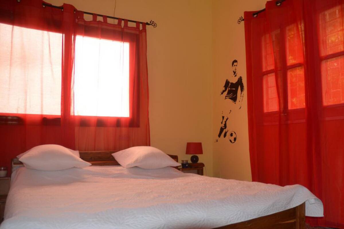 A bedroom with a wooden bed, two white pillows, a nightstand with a lamp, and a window with red curtains.