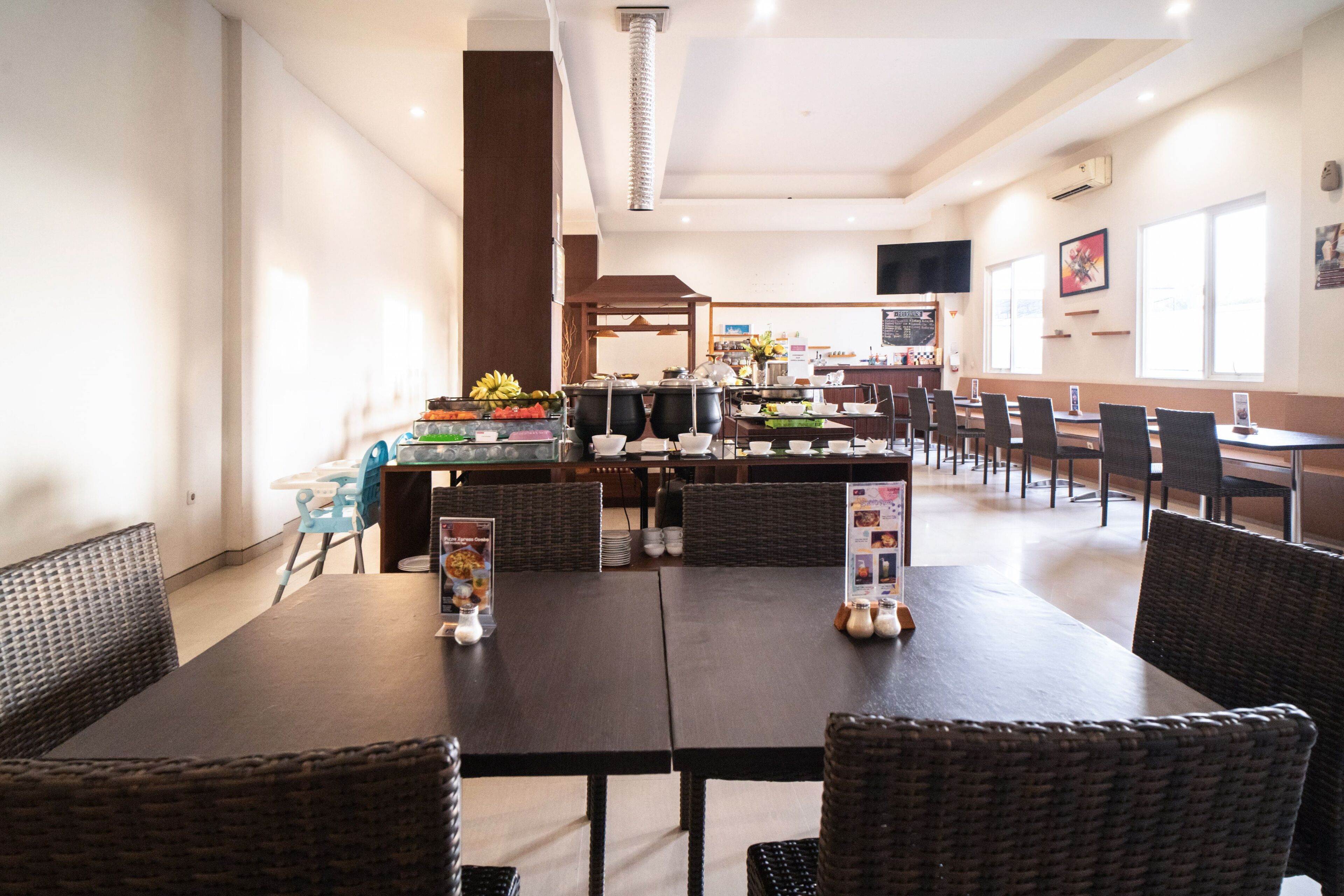 A restaurant with dark wooden tables and wicker chairs, a buffet area with food and drinks, and a television mounted on the wall.