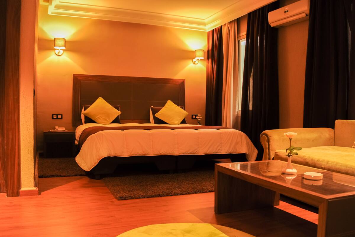 Una habitación de hotel con una cama grande, dos lámparas de noche, una mesita de noche, una mesa de centro de madera y un jarrón con una flor.