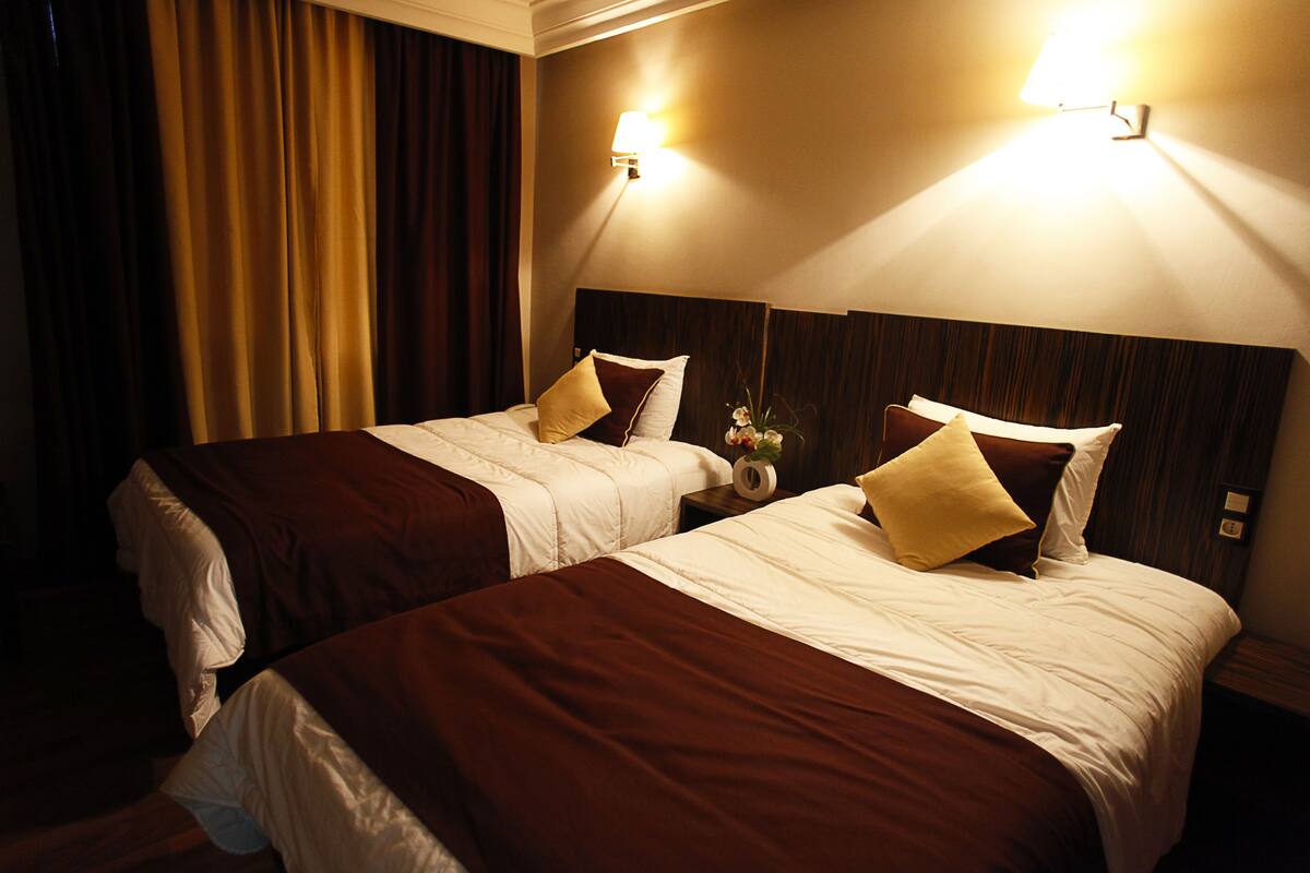 Habitación de hotel con dos camas, cada una con colcha marrón y blanca, cabecero y mesita de noche con lámpara.