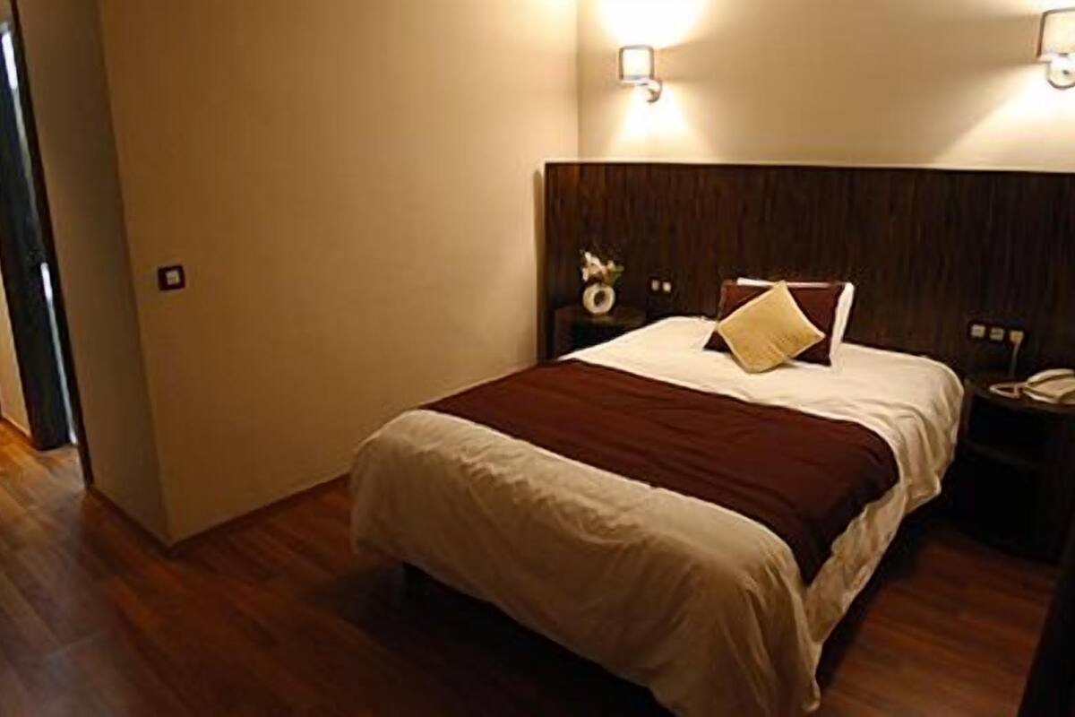 Una cama bien hecha con una colcha marrón y blanca, dos lámparas de noche y una mesita de noche con un teléfono.
