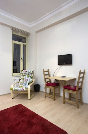 A small room with a wooden floor, a red rug, a wooden chair with a floral cushion, a wooden table with a chair, and a TV mounted on the wall. - Golden Galata Hotel (Istanbul)