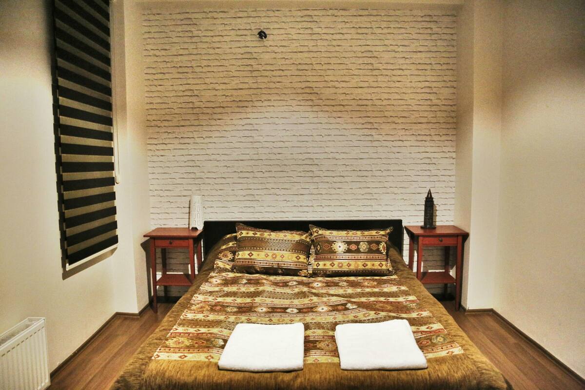 A bedroom with a brick-patterned headboard, a wooden bed, two bedside tables, and a window with blinds.