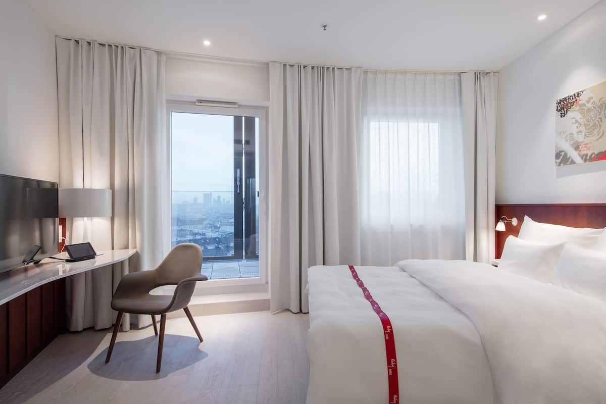 A hotel room with a bed, a desk with a laptop, a chair, a TV, and a large window with curtains.
