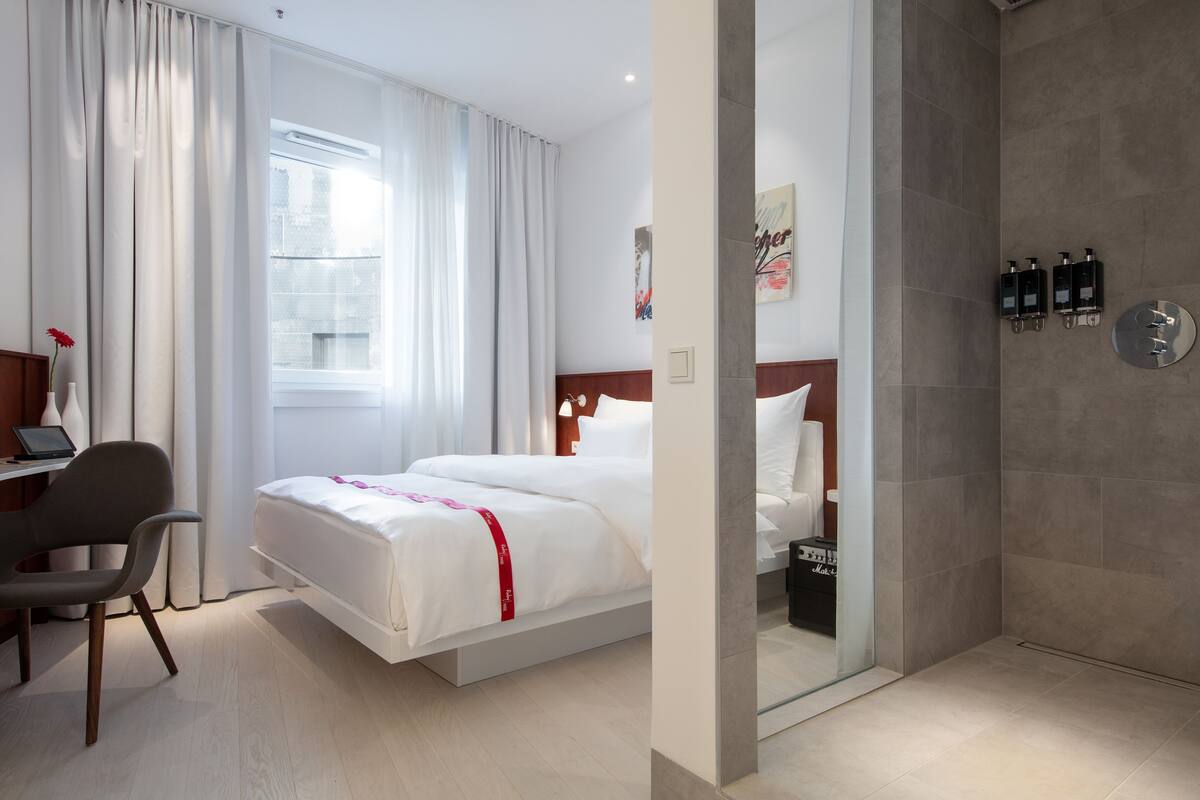 A hotel room with a bed, a desk with a laptop, a chair, a shower area, and a view of the city.