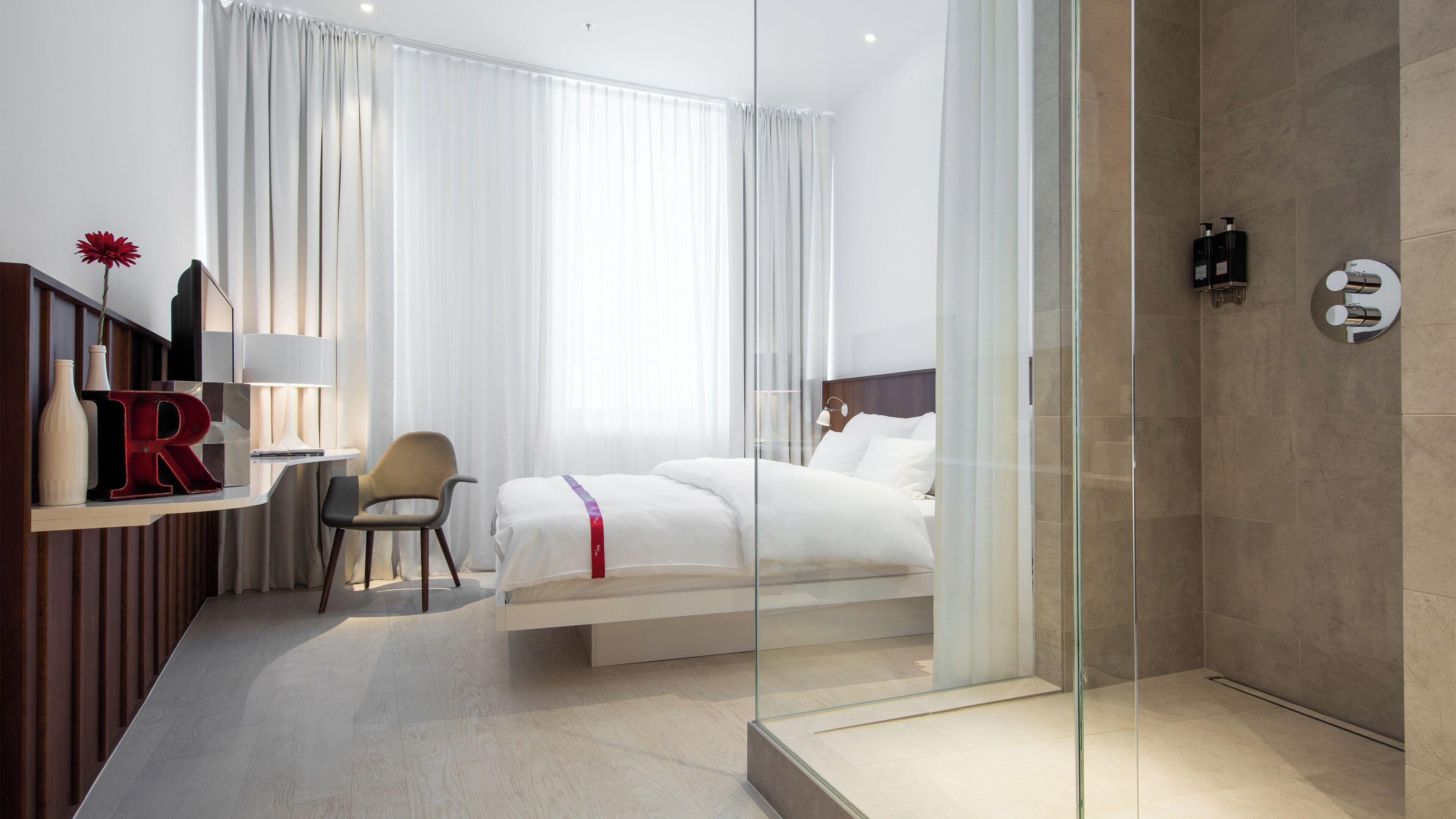 Habitació d'hotel amb llit, escriptori amb cadira, boxa de dutxa de vidre i finestra gran amb cortines.