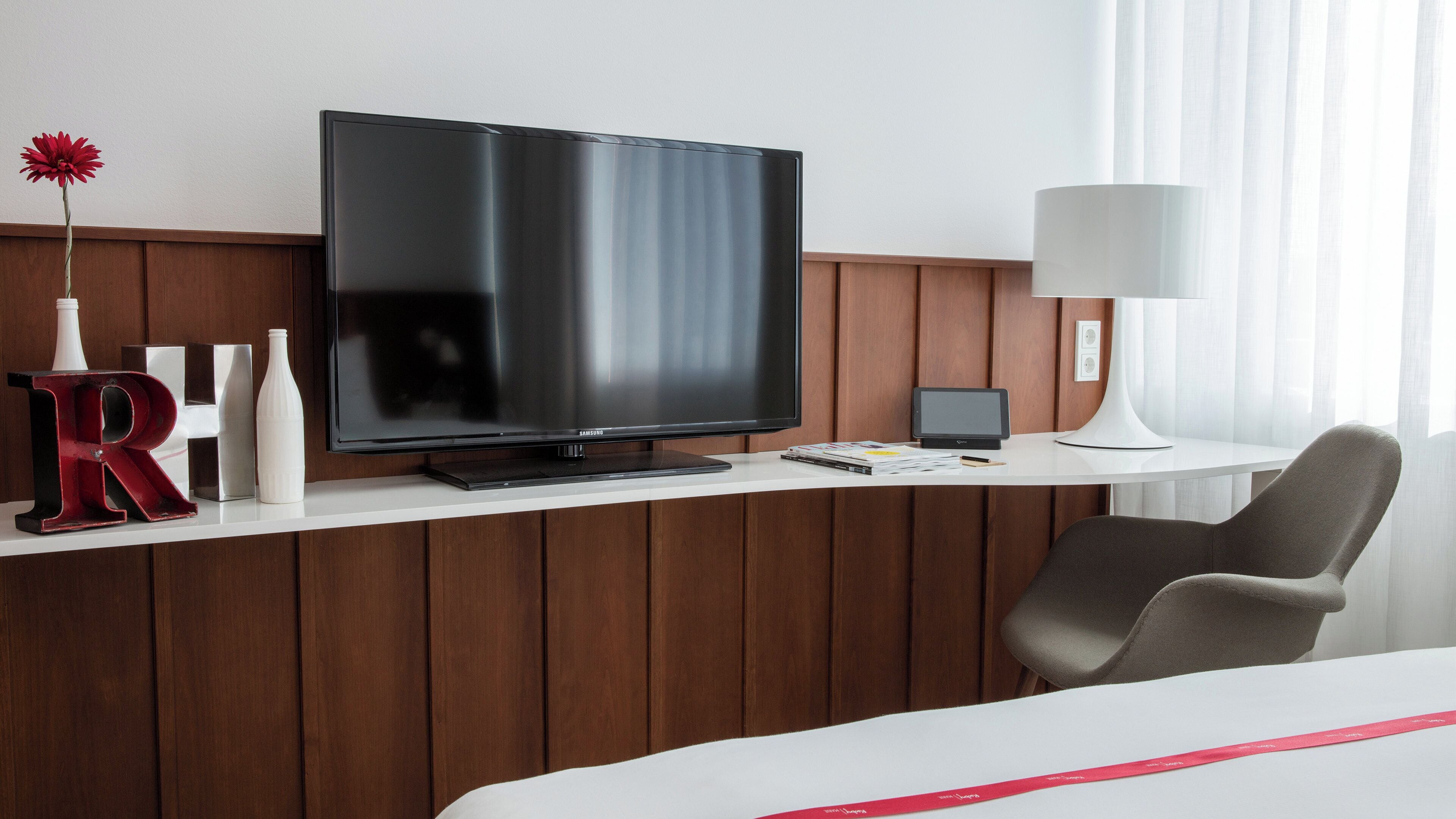 Une chambre d’hôtel moderne équipée d’une télévision à écran plat, d’un bureau avec une lampe, d’une chaise et d’un lit.