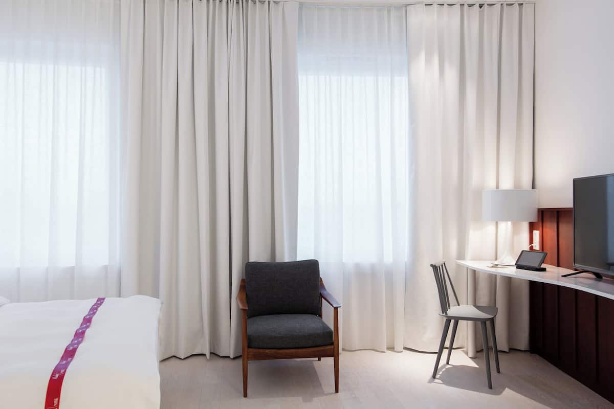 A hotel room with a bed, a desk with a TV, a chair, and large windows with curtains.