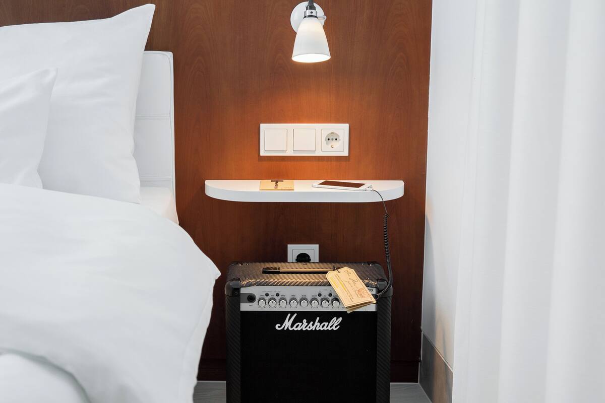 A hotel room with a bed, a nightstand with a lamp, a Marshall amplifier, and a power outlet.