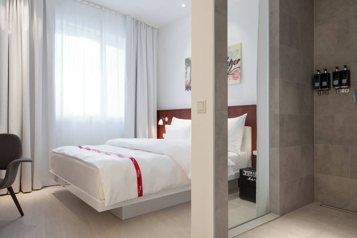 A hotel room with a bed, a desk with a laptop, a chair, a shower area, and toiletries on the wall.