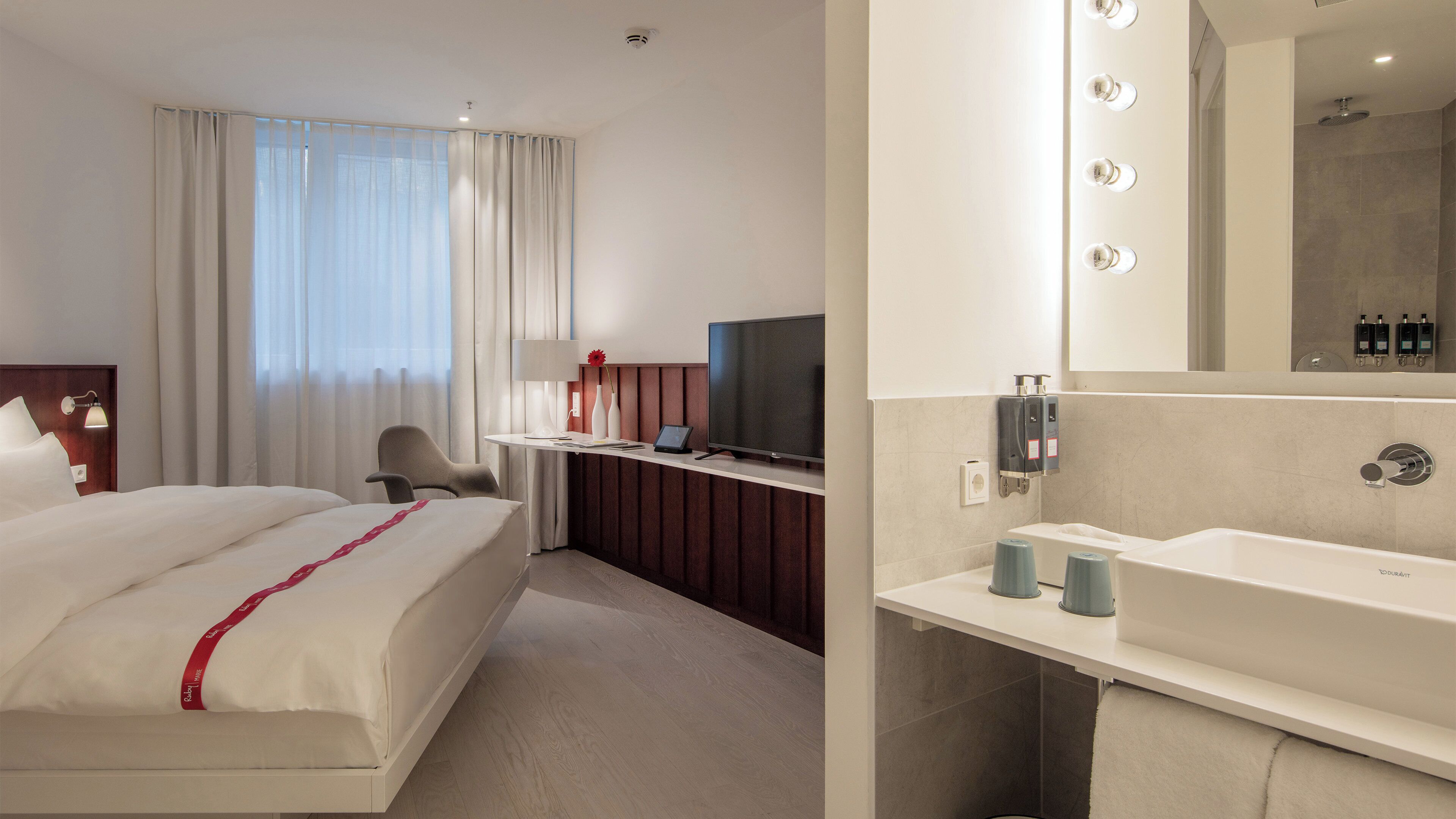 A hotel room with a bed, a desk with a TV, a mirror, and a bathroom with a sink and shower.