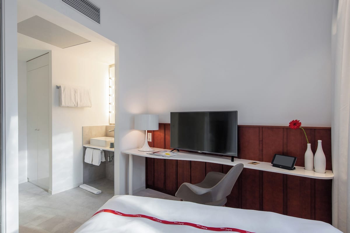 A hotel room with a bed, a TV, a desk with a computer, a chair, and a bathroom visible through an open door.