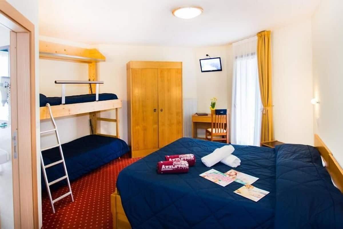 Een kleine hotelkamer met stapelbedden, een houten garderobe, een bureau met stoel, en een klein raam met gordijnen.