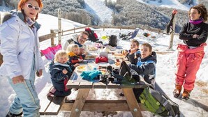 Een groep mensen dineert buiten aan een houten tafel, omringd door met sneeuw bedekte heuvels.