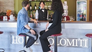 Een barruimte met een barman, een man en een vrouw die zitten. De bar heeft een groene muur met flessen en glazen op planken. Er staat een bord met de tekst 'HOTEL LOMITI'.