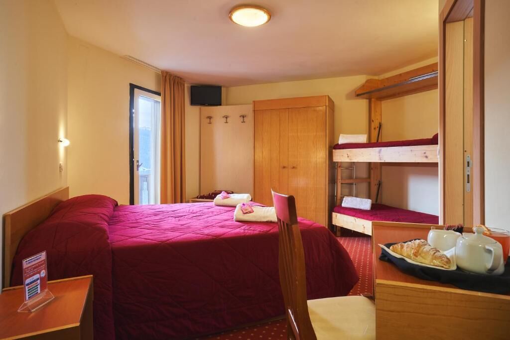 Ein Hotelzimmer mit einem großen Bett, einem kleineren Bett, einem Holzschrank, einem Schreibtisch mit einer Platte Croissants und einem an der Wand montierten Fernseher.