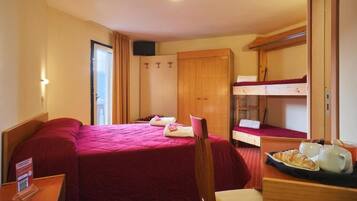 Ein Hotelzimmer mit einem großen Bett, einem kleineren Bett, einem Holzschrank, einem Schreibtisch mit einer Platte Croissants und einem an der Wand montierten Fernseher.