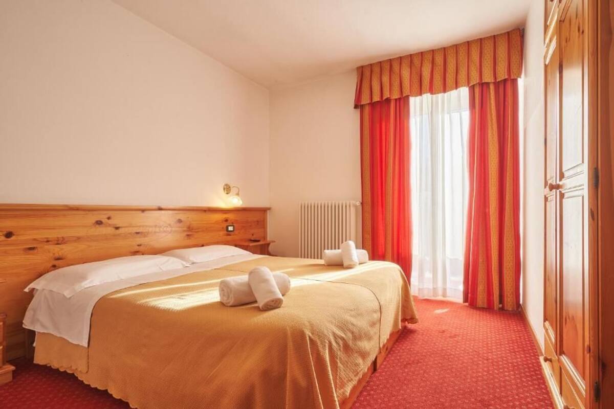 Hotelkamer met een houten hoofdeinde, een tweepersoonsbed met witte lakens, twee opgerolde handdoeken, een radiator en een raam met rode gordijnen.