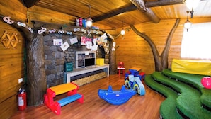 Interior acolhedor de uma cabana de tronco com lareira de pedra, bancos de madeira e área de recreação com um escorrega azul e brinquedos infláveis.
