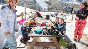 Um grupo de pessoas jantando ao ar livre em uma mesa de madeira, rodeado por colinas cobertas de neve.