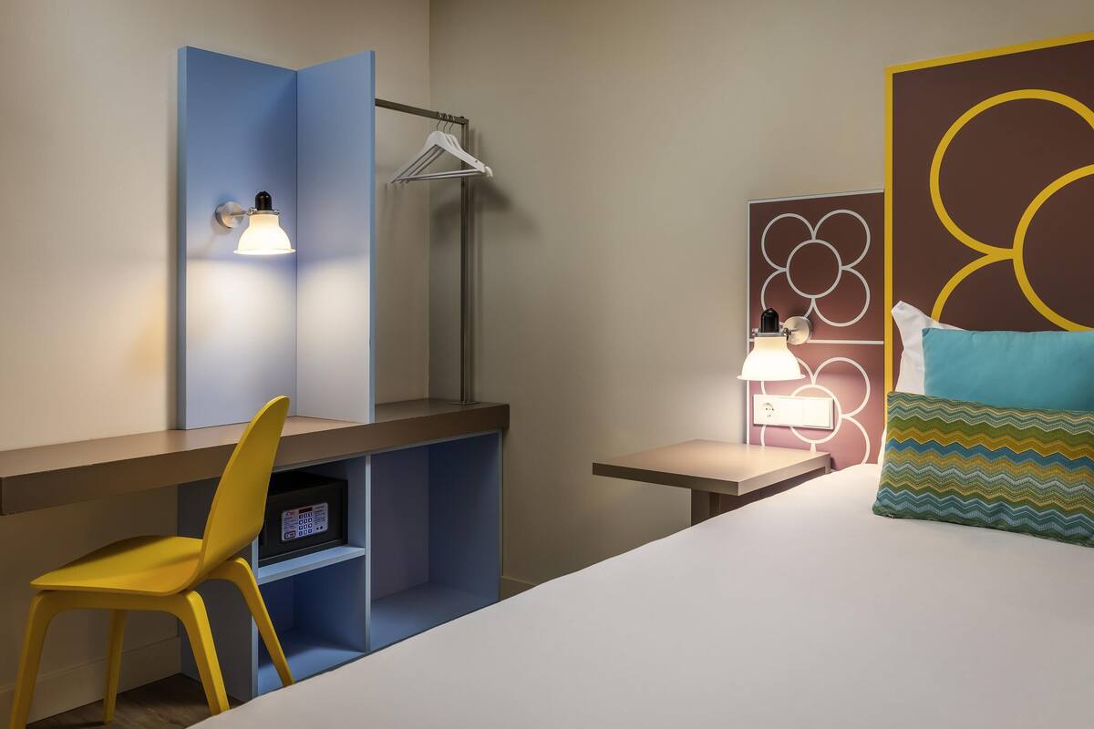 A hotel room with a bed, a desk with a chair, a desk lamp, a wall-mounted shelf, and a wall-mounted light fixture.