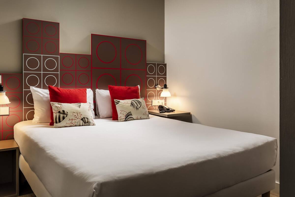 A hotel room with a large bed, a bedside table with a lamp, and a wall with geometric patterns.