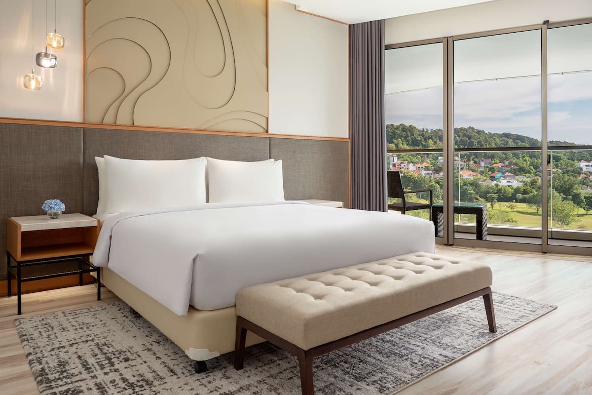 Ein modernes Hotelzimmer mit einem großen Bett, einer Bank, einem Nachttisch und Blick auf eine grüne Landschaft durch große Fenster.