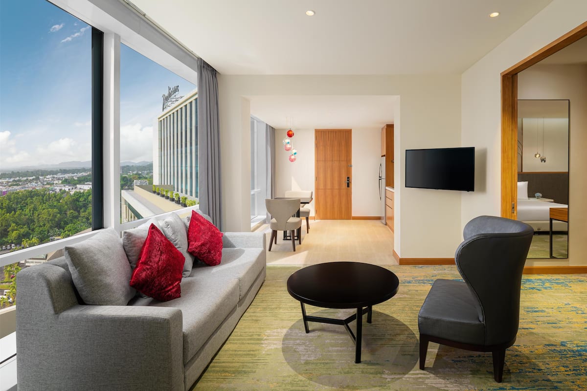 Ein modernes Hotelzimmer mit großem Fenster und Blick auf die Stadt, einer Couch mit roten Kissen, einem runden Couchtisch, einem grauen Sessel und einem an der Wand befestigten Flachbildfernseher.